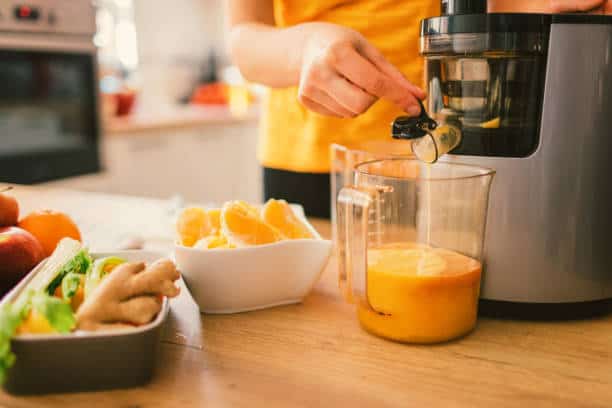 Person using a juicer to make fresh orange juice on a kitchen counter with ingredients including oranges, ginger, and vegetables nearby.