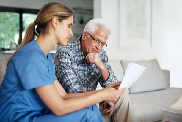 A nurse in blue scrubs shows documents to an elderly man with glasses and gray hair sitting on a couch.