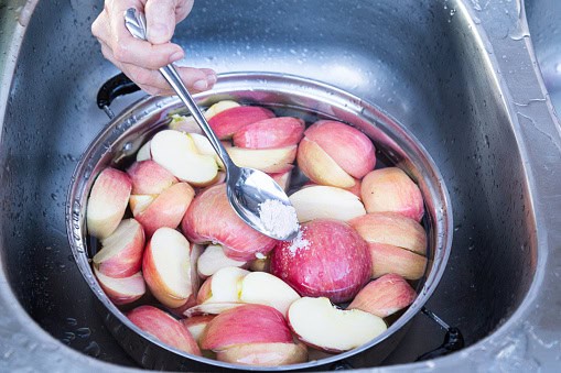 A hand is adding a spoonful of white powder to a metal bowl filled with halved apples submerged in water in a sink.