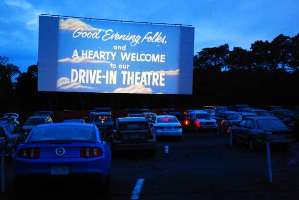 A drive-in theater with several cars parked facing a large screen displaying a welcome message against a dusky sky.