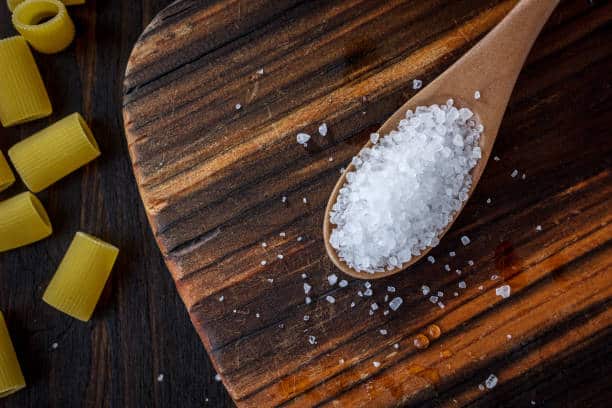 A wooden spoon filled with coarse salt rests on a wooden cutting board, with uncooked pasta scattered nearby.