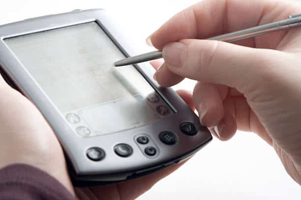 A person uses a stylus to interact with an early model PDA with buttons on the bottom and a grayscale touch screen.