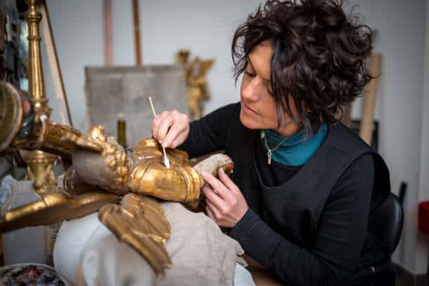 A person with curly hair meticulously cleans a golden statue using a small brush while seated in a workshop.