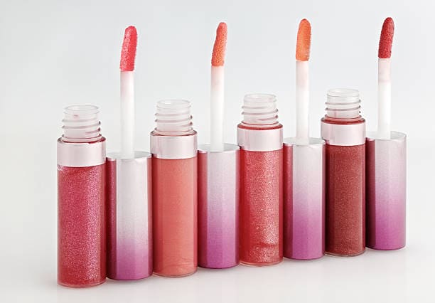 Six lip gloss tubes with different shades of pink and orange are lined up, each with an applicator wand inserted. The background is plain white.