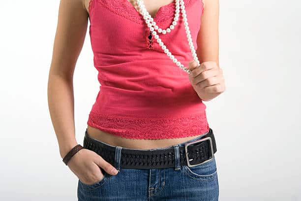 A person wearing a pink tank top, jeans, and a black belt holds a pearl necklace against a plain white background.