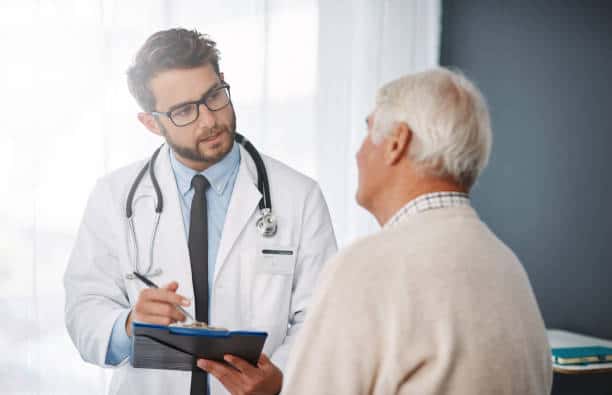 A doctor wearing a white coat and stethoscope holds a clipboard while talking to an elderly man in a sweater.