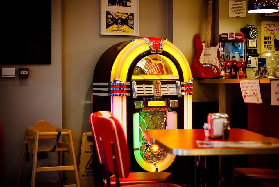 A brightly lit jukebox stands against the wall of a retro-themed diner, surrounded by vintage decor, a guitar on the wall, and a table with red chairs in the foreground.