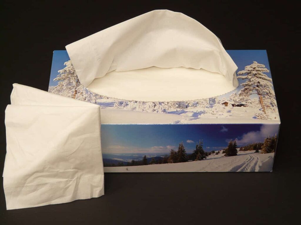 A rectangular box of tissues with a winter landscape design, one tissue partially pulled out and another tissue placed beside the box on a dark surface.