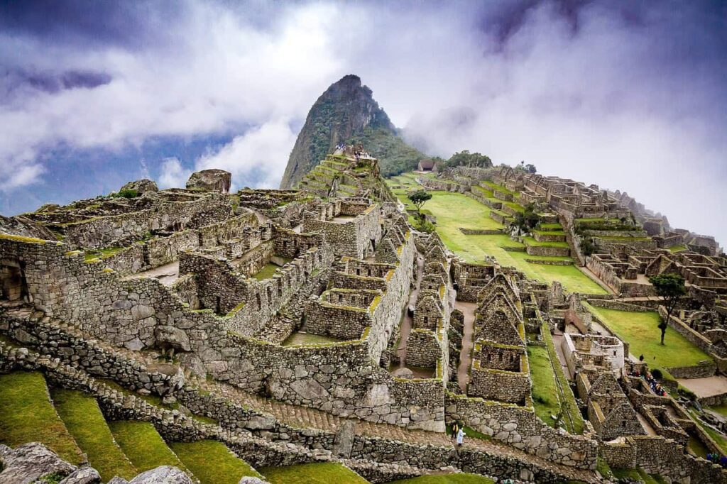 A view of Machu Picchu's stone ruins with terraces and green grass, set against a backdrop of misty mountains and cloudy sky.