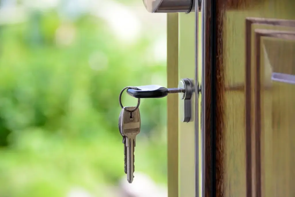 Close-up of a set of keys inserted in the lock of an open wooden door with a blurred green background outside.