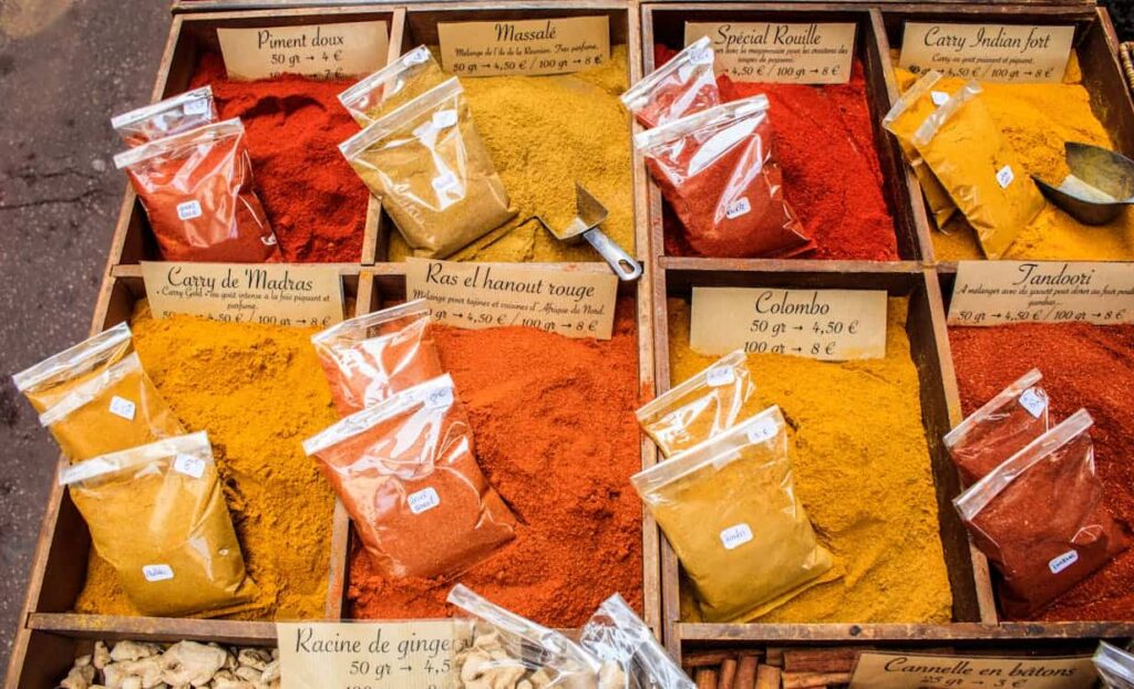 Various types of spices and seasonings displayed in open containers and plastic bags at a market, each labeled with names and prices.