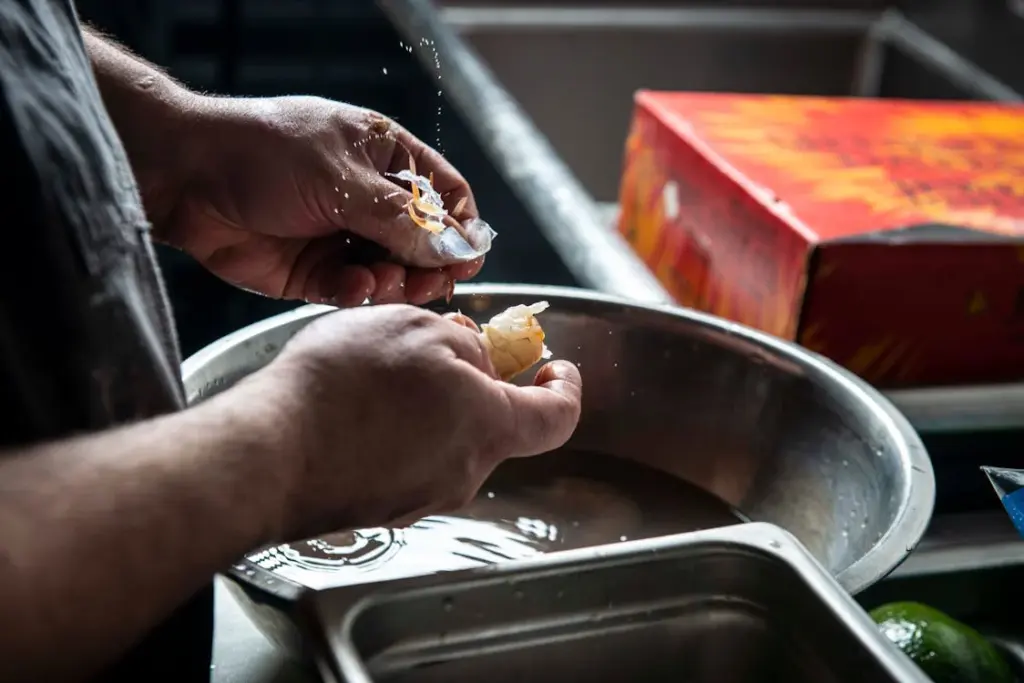 A person is peeling shrimp over a stainless steel bowl, with a partially opened red box nearby.