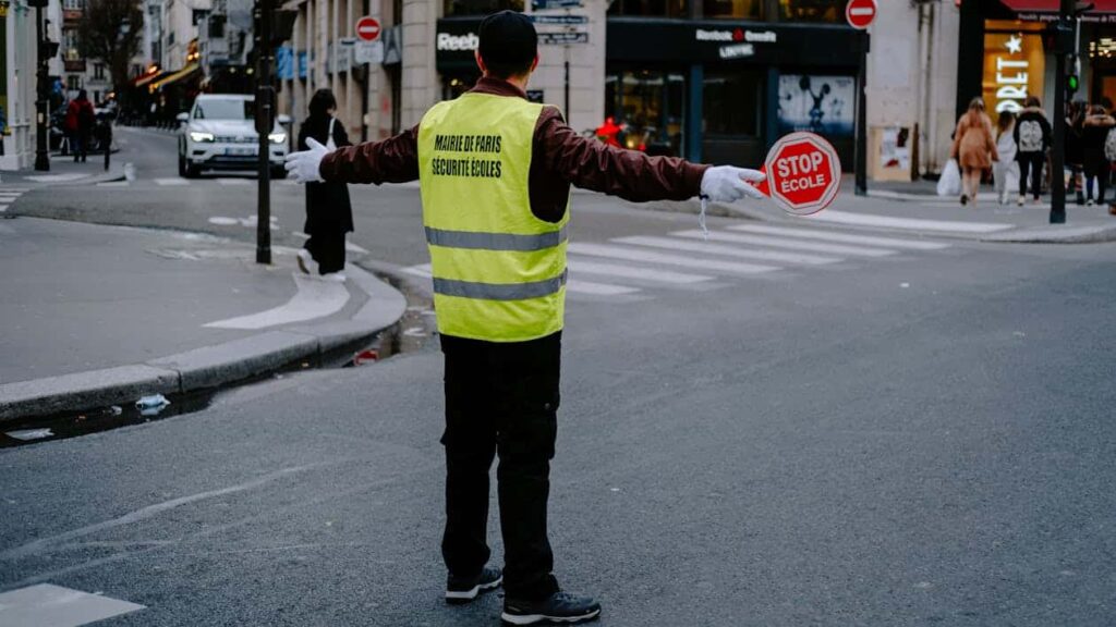 A crossing guard wearing a yellow vest holds a stop sign in the middle of a crosswalk, stopping traffic as pedestrians cross the street in an urban area.