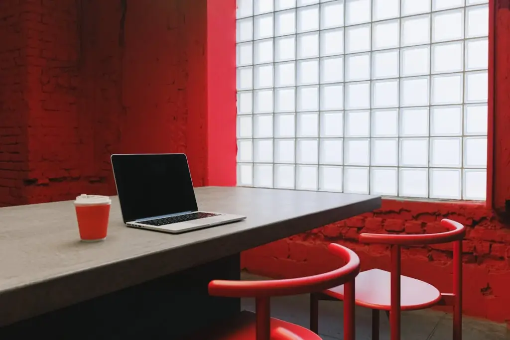 A laptop and coffee cup on a table in a room with red walls and chairs, illuminated by a frosted glass window.
