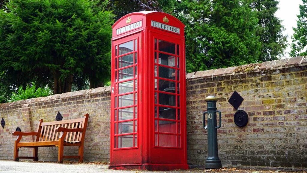 A red telephone booth stands next to a wooden bench and a brick wall, with green trees in the background.