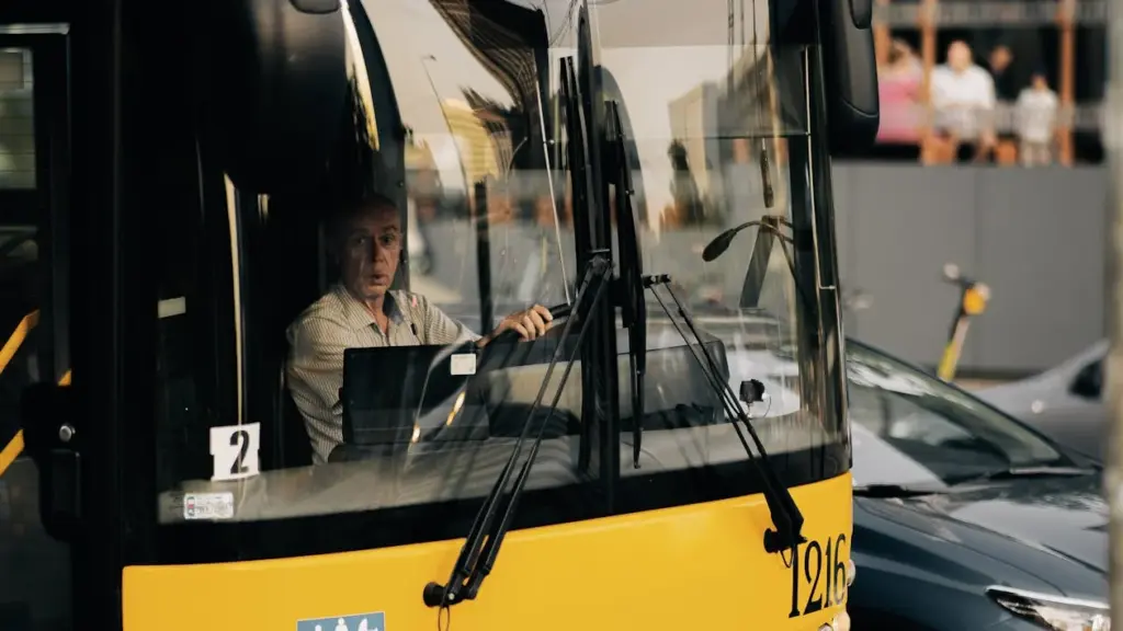 A bus driver sits at the wheel of a yellow bus in traffic.