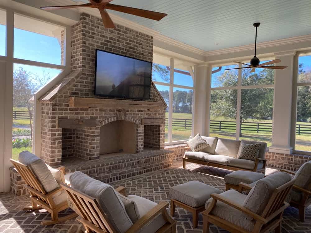 A sunlit enclosed porch features a brick fireplace with a TV mounted above, surrounded by cushioned wooden furniture, large windows, and ceiling fans.