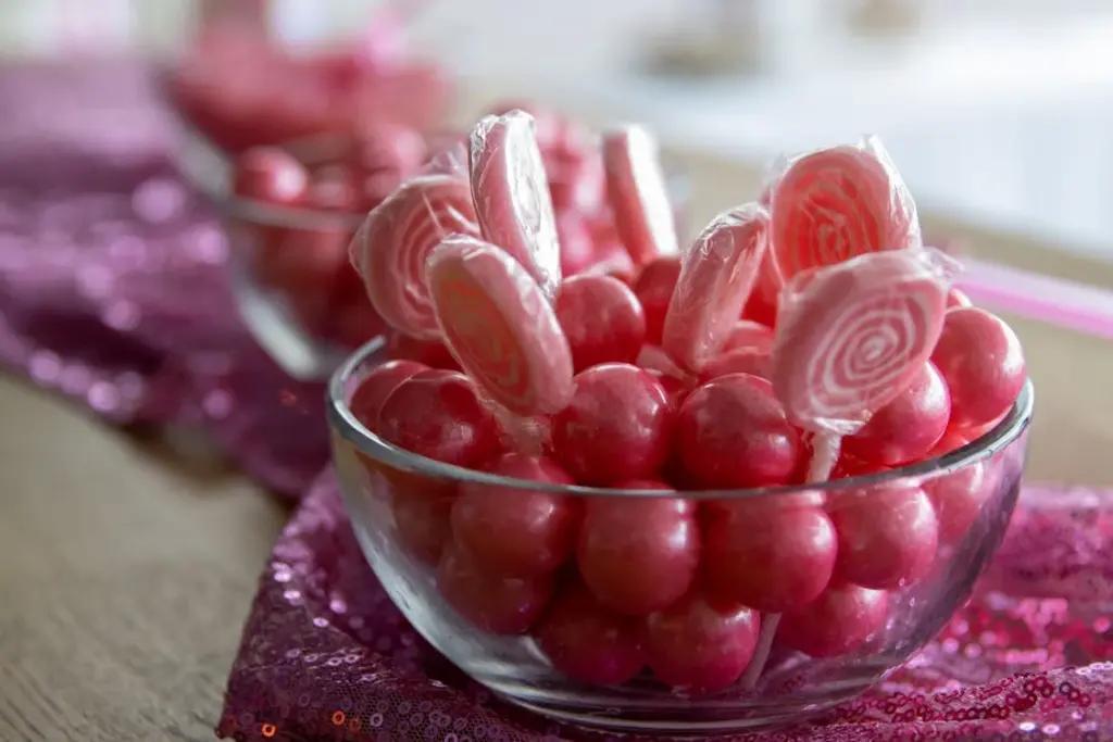 Glass bowls filled with pink gumballs and swirl lollipops, placed on a sequined pink fabric.