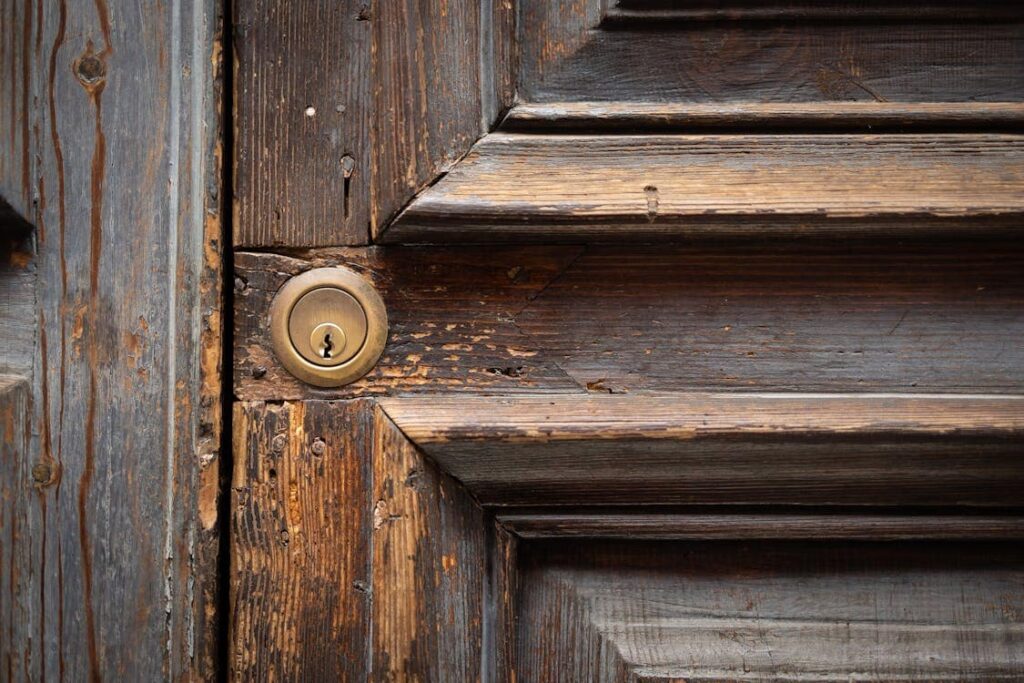 Close-up of an aged wooden door with visible textures and a brass keyhole on the left side. The wood shows signs of wear and weathering.