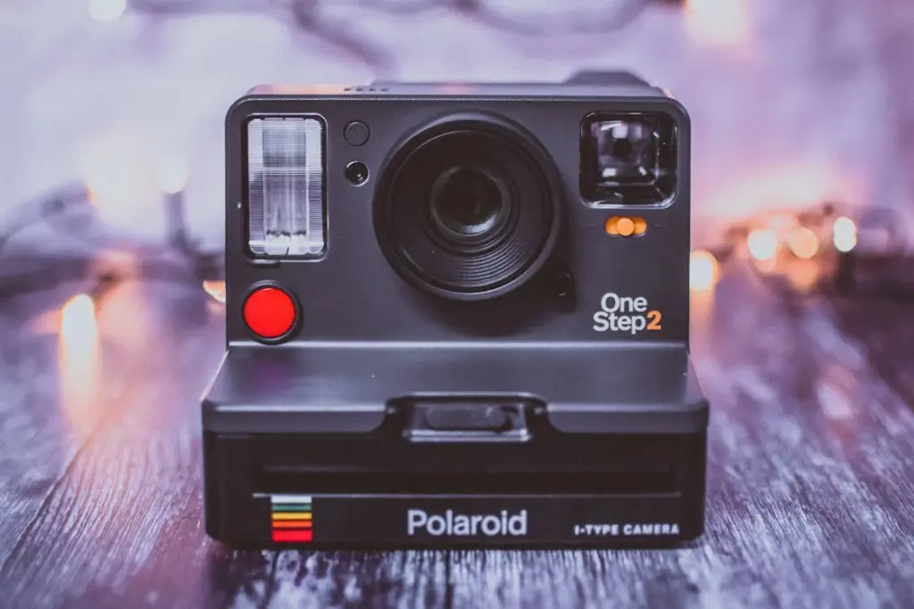 A Polaroid OneStep2 camera is placed on a table with a blurred background of string lights. The camera has a black finish with a red button and reflective elements.