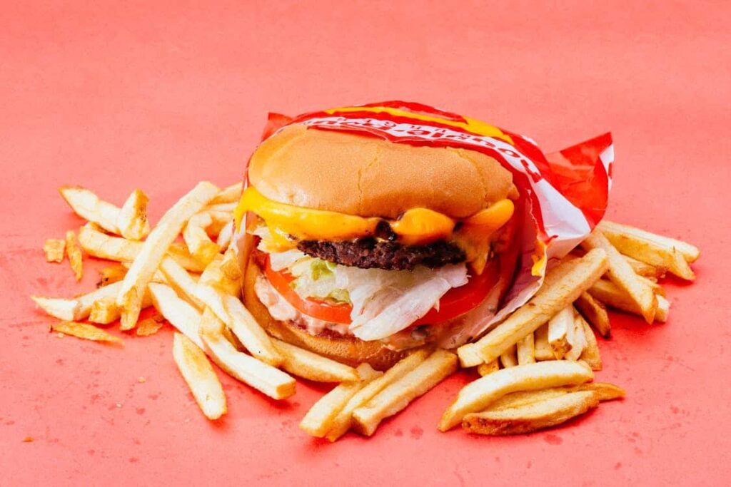 A cheeseburger with lettuce, tomato, and a beef patty partially wrapped in paper, surrounded by French fries on a red background.