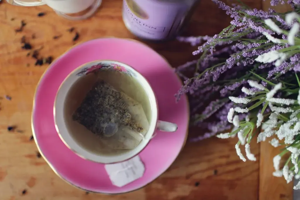 A teacup sits on a pink saucer, containing a tea bag steeping in hot water. There is a jar and a bundle of lavender flowers on the wooden table.