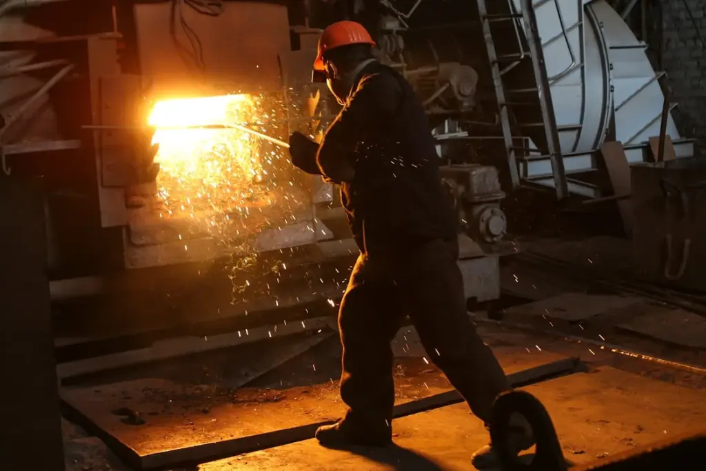 A worker in protective gear operates equipment near molten metal, producing sparks in an industrial setting.