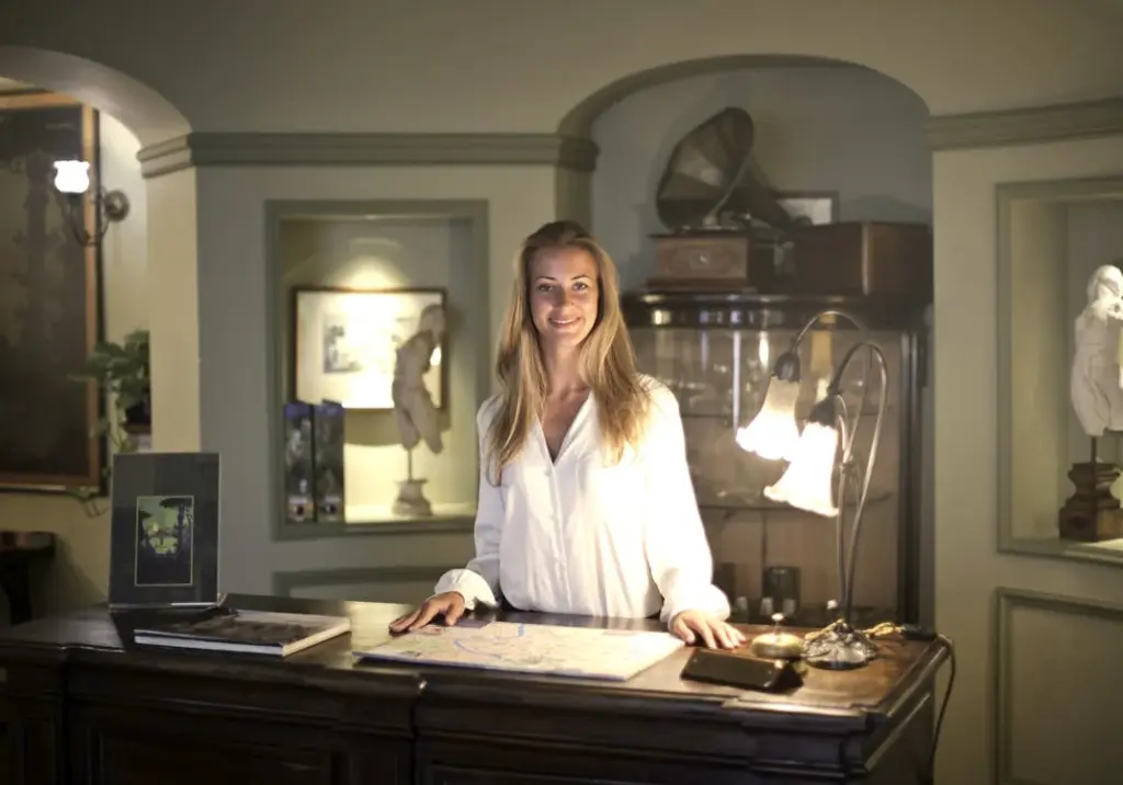 A woman in a white blouse stands behind a wooden desk with various items, including a map and a lamp, in an elegantly decorated room with framed artwork and sculptures.