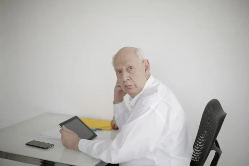 An older man in a white shirt sits at a white desk, holding a tablet in one hand and looking at the camera. A smartphone and papers are also on the desk.