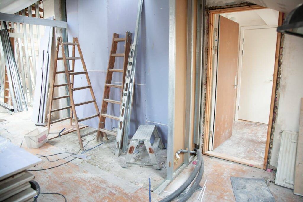 A room under construction with ladders, a step stool, and various construction materials against an unfinished wall. An open door leads to another room.