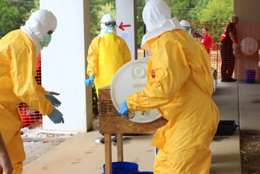 Several individuals wearing yellow hazmat suits work with a large white barrel outside, with red safety fencing and greenery in the background.