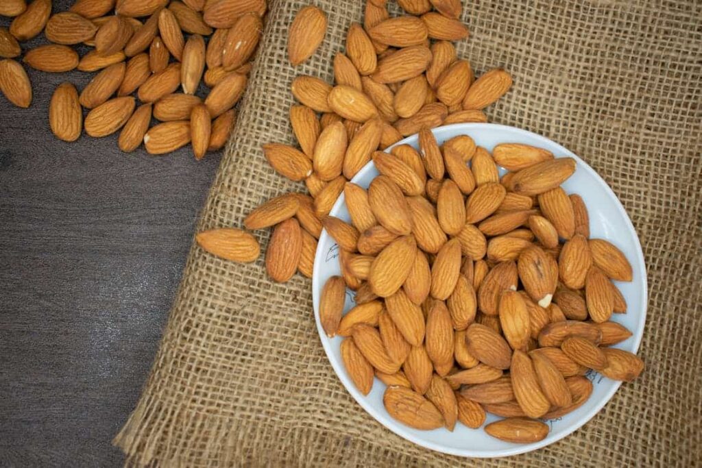 A white bowl filled with almonds on a burlap cloth, with some almonds scattered around it and on a dark wooden surface.