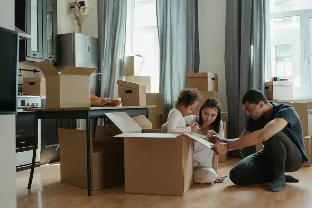 A couple and a child unpack boxes in a kitchen while moving into a new home, with cardboard boxes scattered around.