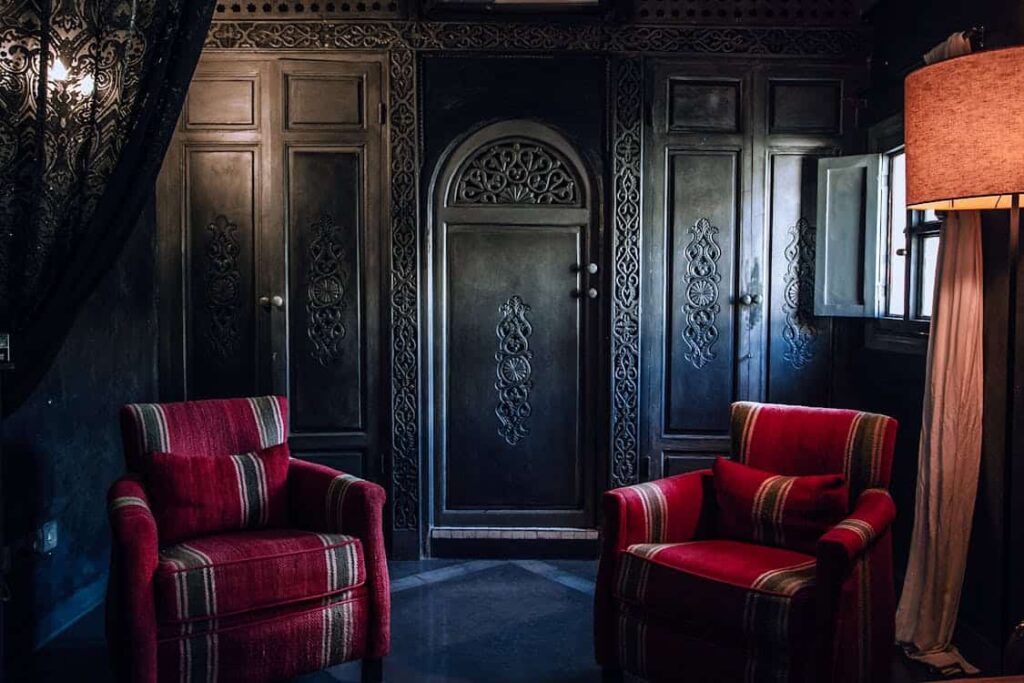 Two red armchairs with striped patterns facing each other in a dimly lit room with ornate black wooden doors and a window partially covered by a curtain.