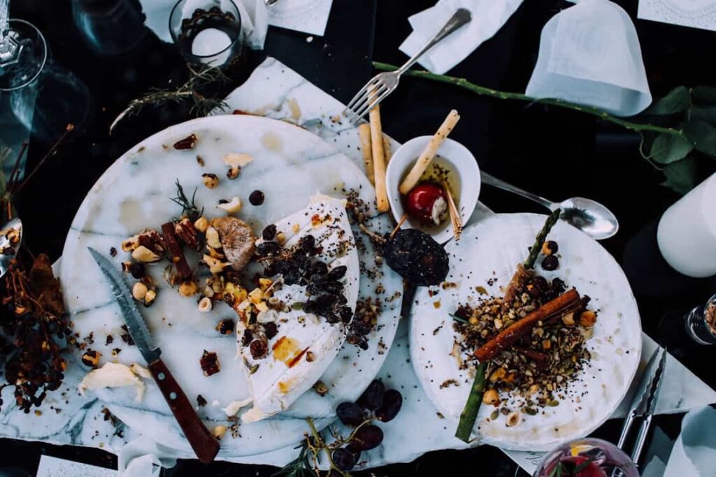 A dining table with marble plates featuring partially eaten food. The table is cluttered with utensils, crumbs, seeds, vegetables, a small bowl with breadsticks and a cherry, and various decorative items.