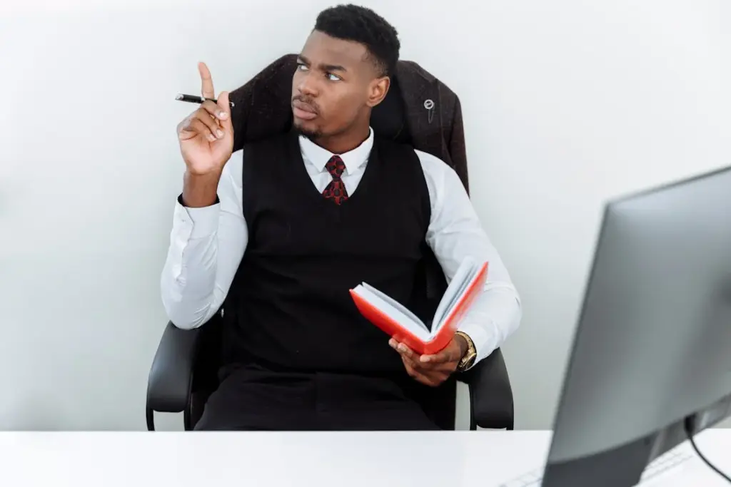 A man in business attire sits at a desk, holding an open red book, and points his finger upward as if making a point or having an idea. A computer monitor is visible in the foreground.