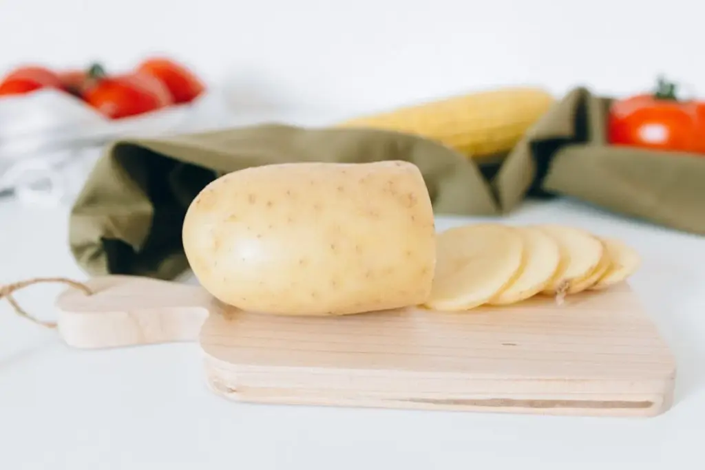 A whole potato and several potato slices are placed on a wooden cutting board, with tomatoes and corn visible in the background.