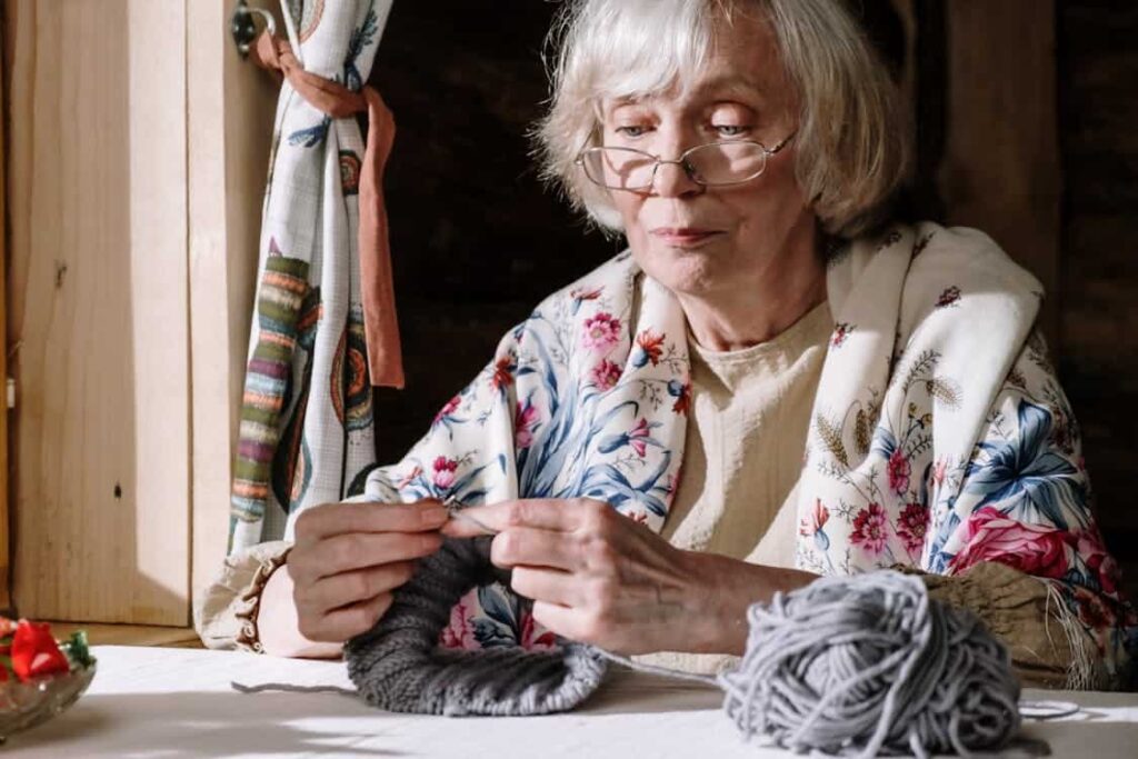 An elderly woman with glasses and a floral shawl is knitting with gray yarn at a wooden table near a window.