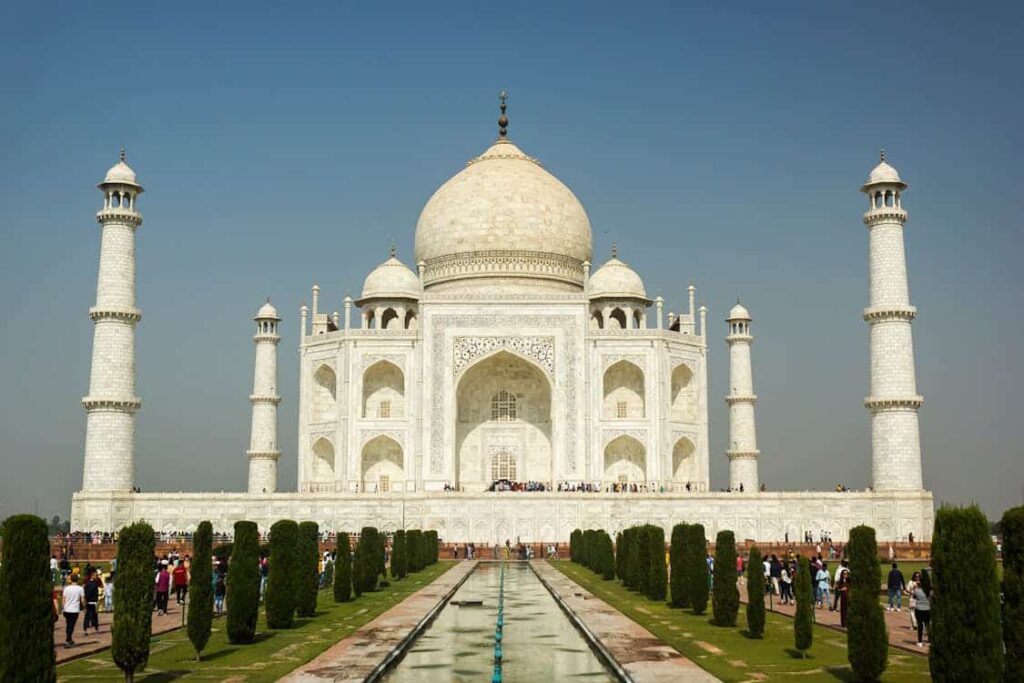 The Taj Mahal, a white marble mausoleum with minarets, reflecting in a pool, with tourists walking in front. Clear blue sky in the background.