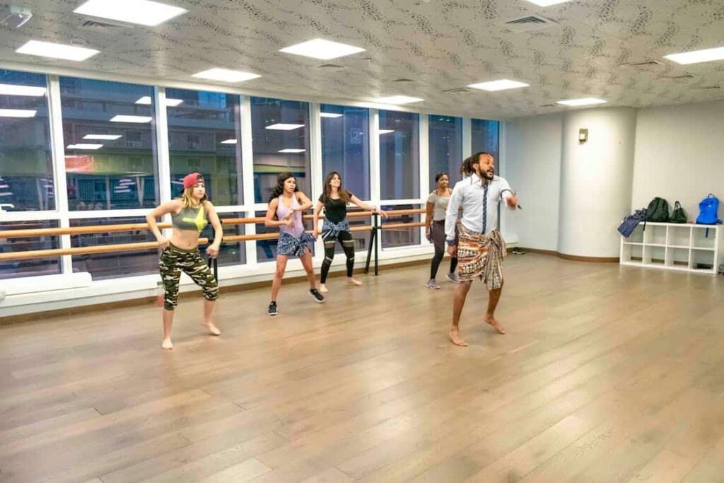 A group of people participates in a dance class in a brightly lit studio with large windows and wooden floors.