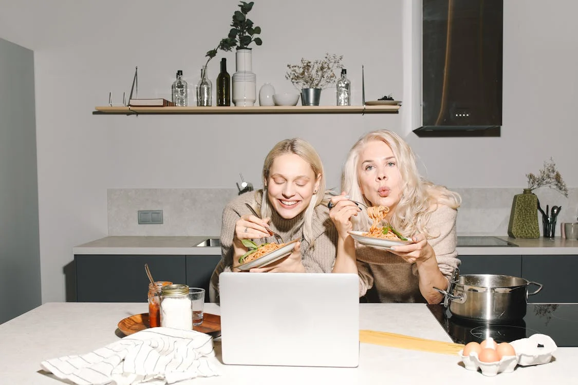 Two people sit in a kitchen eating from bowls of food while looking at a laptop. Various kitchen items are on the countertop, including a jar, a pot, and some eggs.
