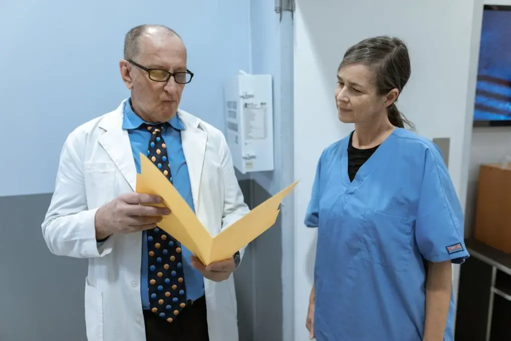 A male doctor reviews a yellow folder while a female nurse in blue scrubs listens attentively in a medical facility.