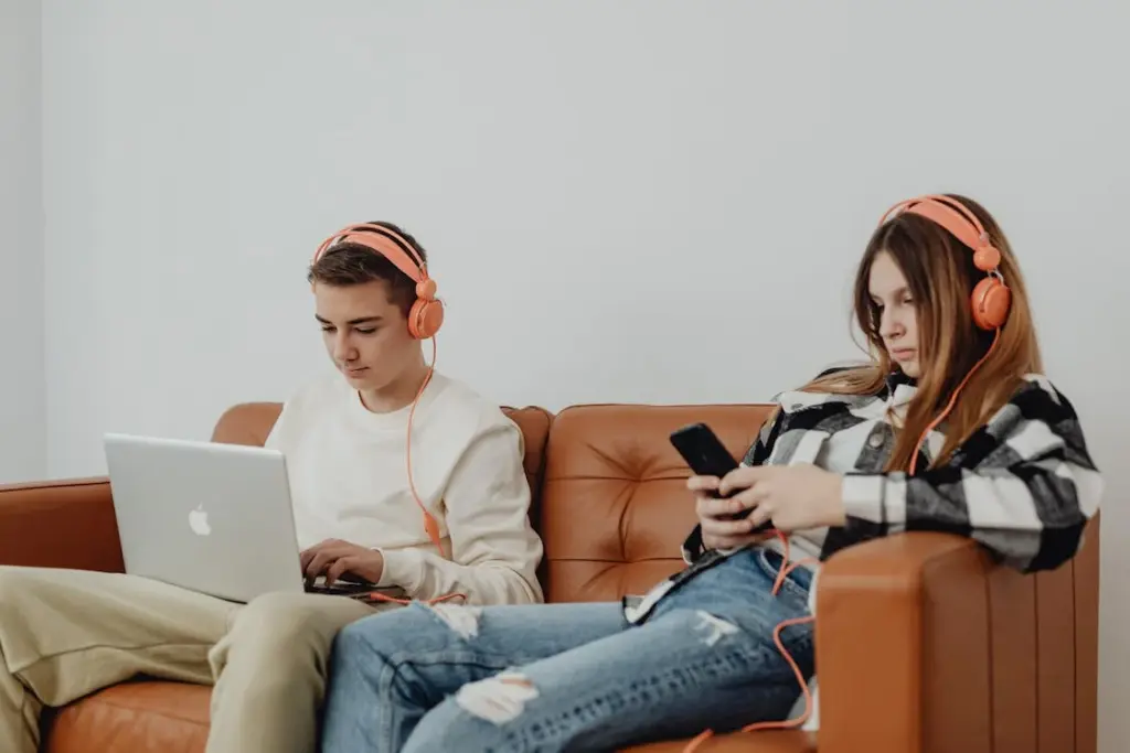 Two individuals seated on a brown couch, both wearing orange headphones; one is using a laptop, and the other is focused on a smartphone.
