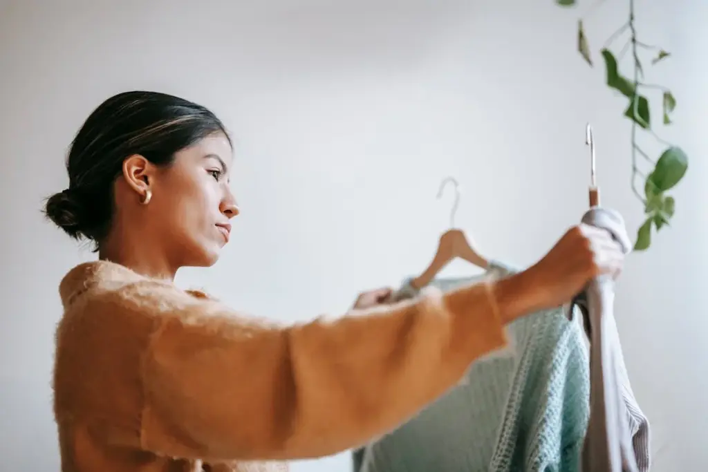 A woman with tied-back hair is holding up two sweaters on hangers, examining them closely. A green plant hangs from the ceiling in the background.