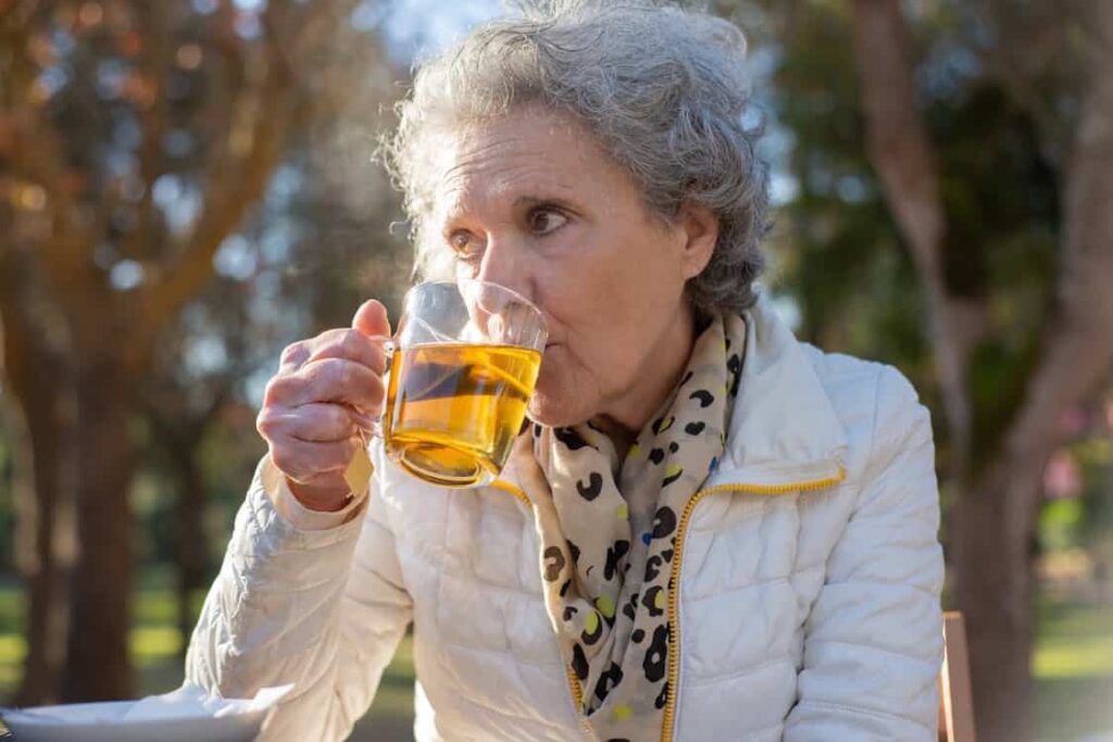 An elderly woman with grey hair sips tea from a glass mug while seated outdoors. She is wearing a white jacket and a light-colored scarf with a pattern. Trees and sunlight are visible in the background.