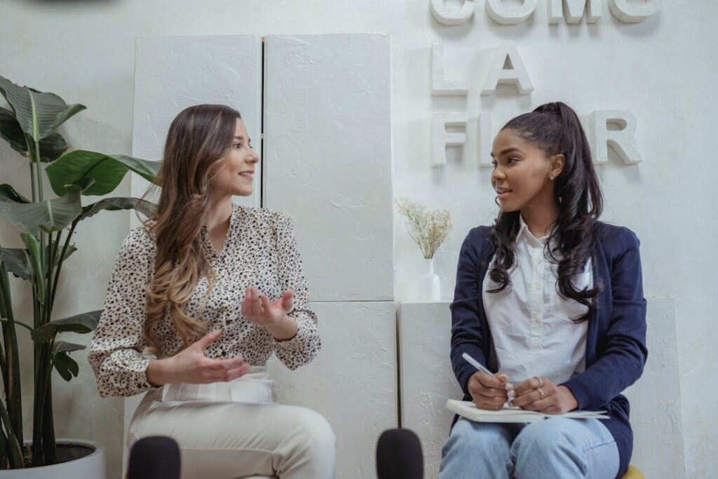 Two women sit and converse in a light-colored room with minimalist decor. One gestures with her hands while speaking, and the other takes notes.