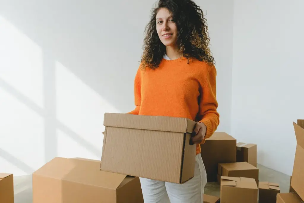 A woman with curly hair wearing an orange sweater holds a cardboard box. She stands among other unpacked boxes in a brightly lit room.