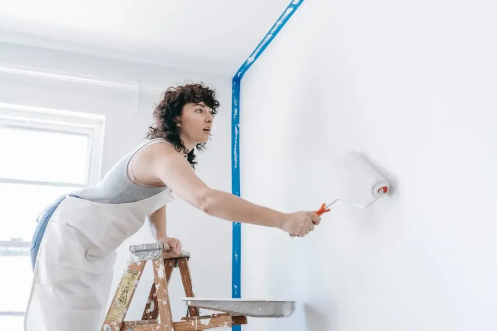 A person standing on a ladder is painting a white wall with a roller. The window is open and a strip of blue painter's tape is visible on the wall.