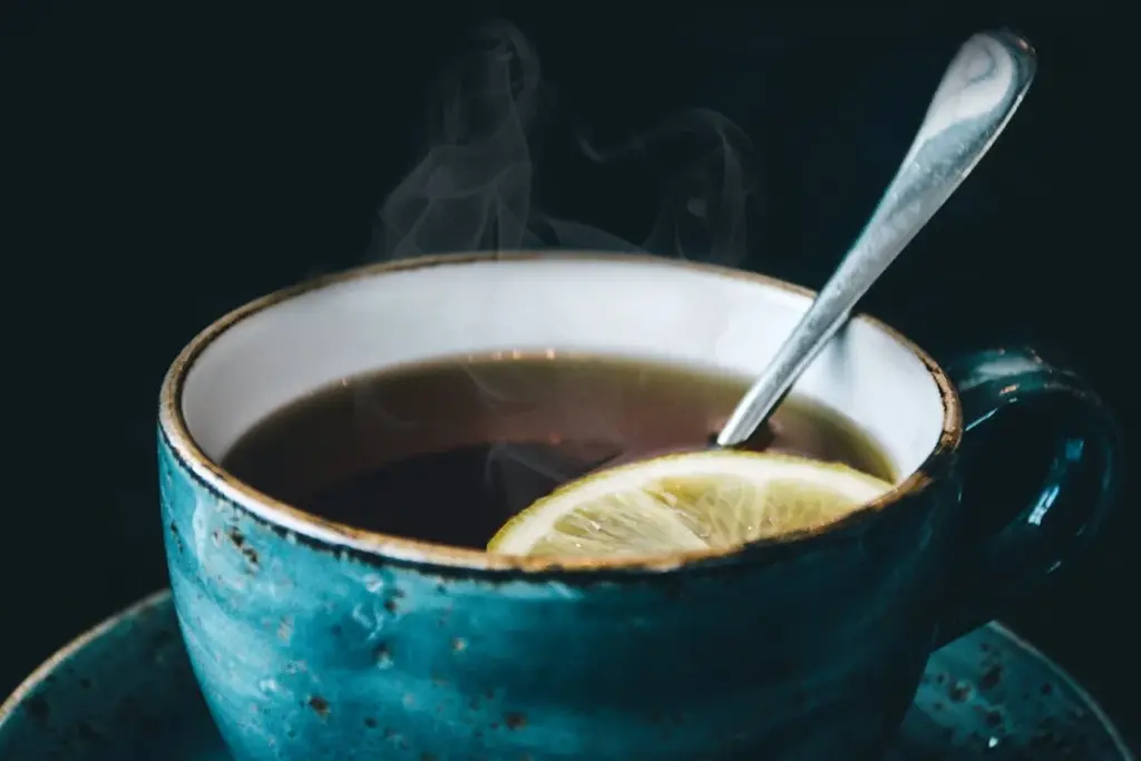 A steaming cup of tea in a blue ceramic cup with a lemon slice and a spoon inside.