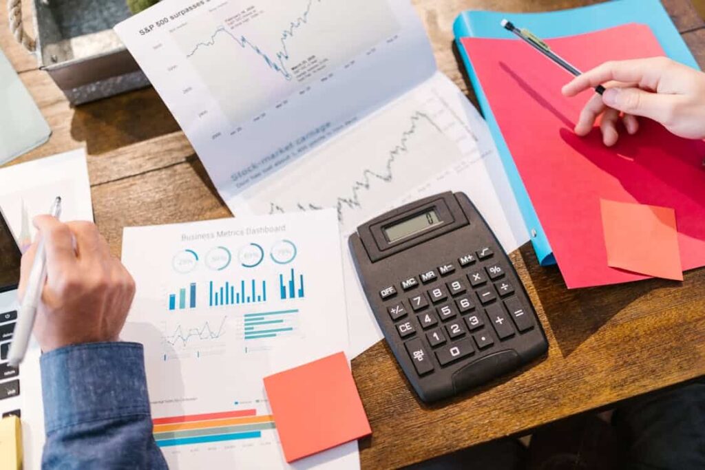 Two people sit at a wooden table analyzing financial charts and graphs. A calculator, red notebook, blue folder, and sticky notes are also on the table.
